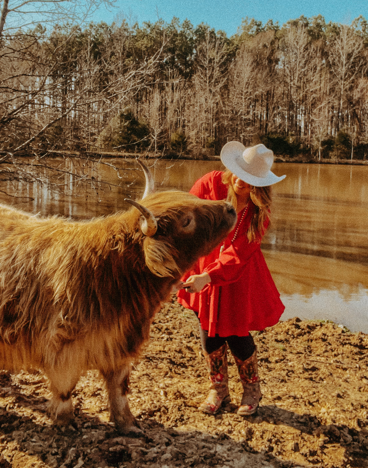 Brooklyn in a red dress and white cowboy hat sharing a sweet moment with Charlsie the highland cow beside the farm pond